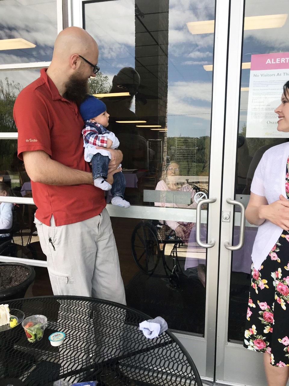man holding a baby in front of a window to say hi to the residents inside the building