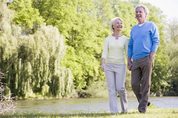 senior woman and senior man walking along the edge of a pond with large green trees in the background