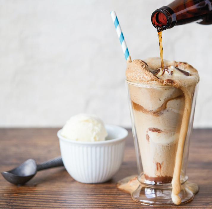 root beer float with a blue and white striped straw placed on tabletop next to bowl of ice cream and ice cream scooper